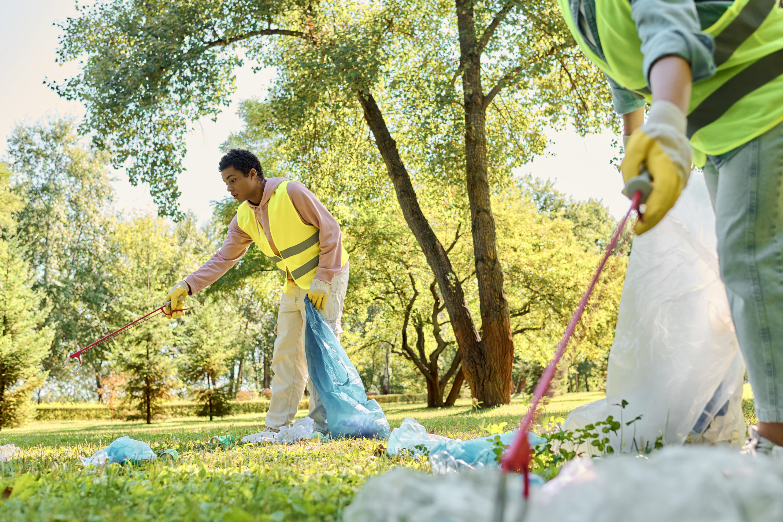 A diverse couple, wearing safety vests and gloves, working together to clean up a grassy park.